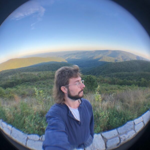 Photo of Andrew Krysinski in a fish eye lens in front of a scenery of mountains and rolling hills