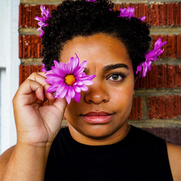 Headshot of Amber Bills with purple flowers in her hair and a purple flower in front of her eye