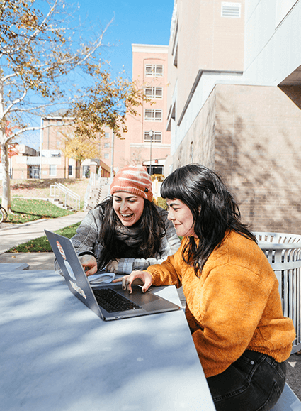 Two students sit at an outdoor table outside the Brandcenter looking at a laptop