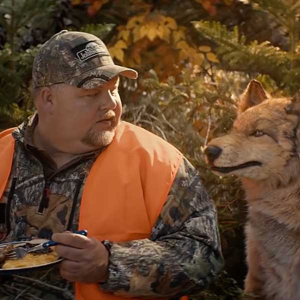 A screengrab from a Johnsonville Sausage commercial with a hunter eating and a fake wolf next to him talking