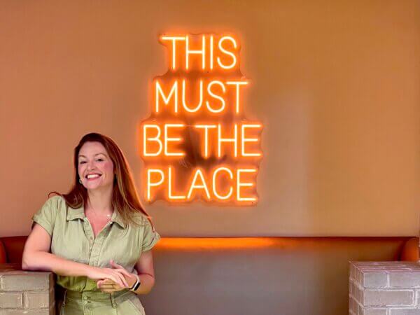 Holly Hessler standing in front of the neon "This Must Be The Place" sign.