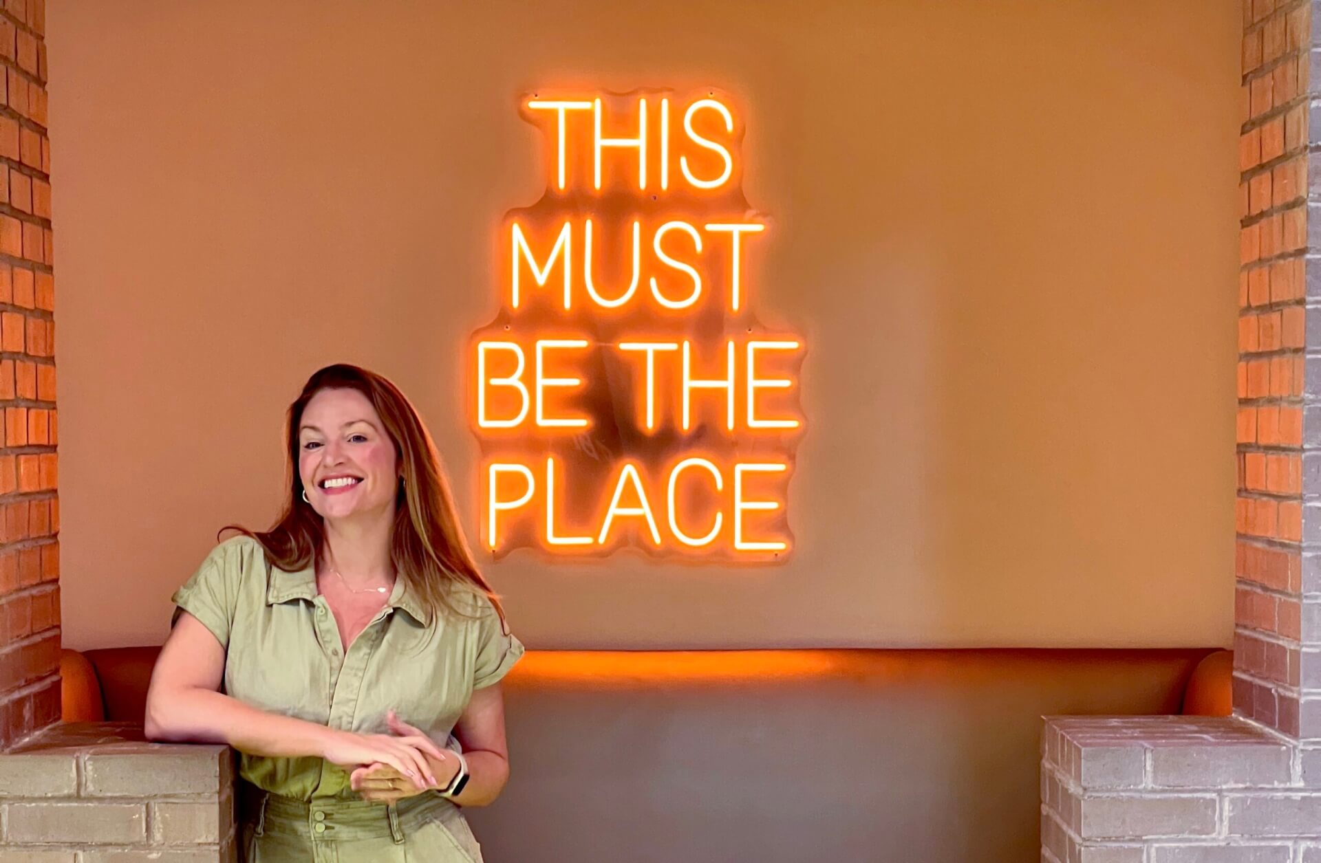 Holly Hessler standing in front of the neon "This Must Be The Place" sign.