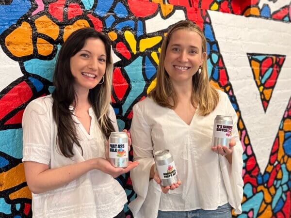 celeste and catherine holding cans of e b t b in front of the brandcenter mural in basement
