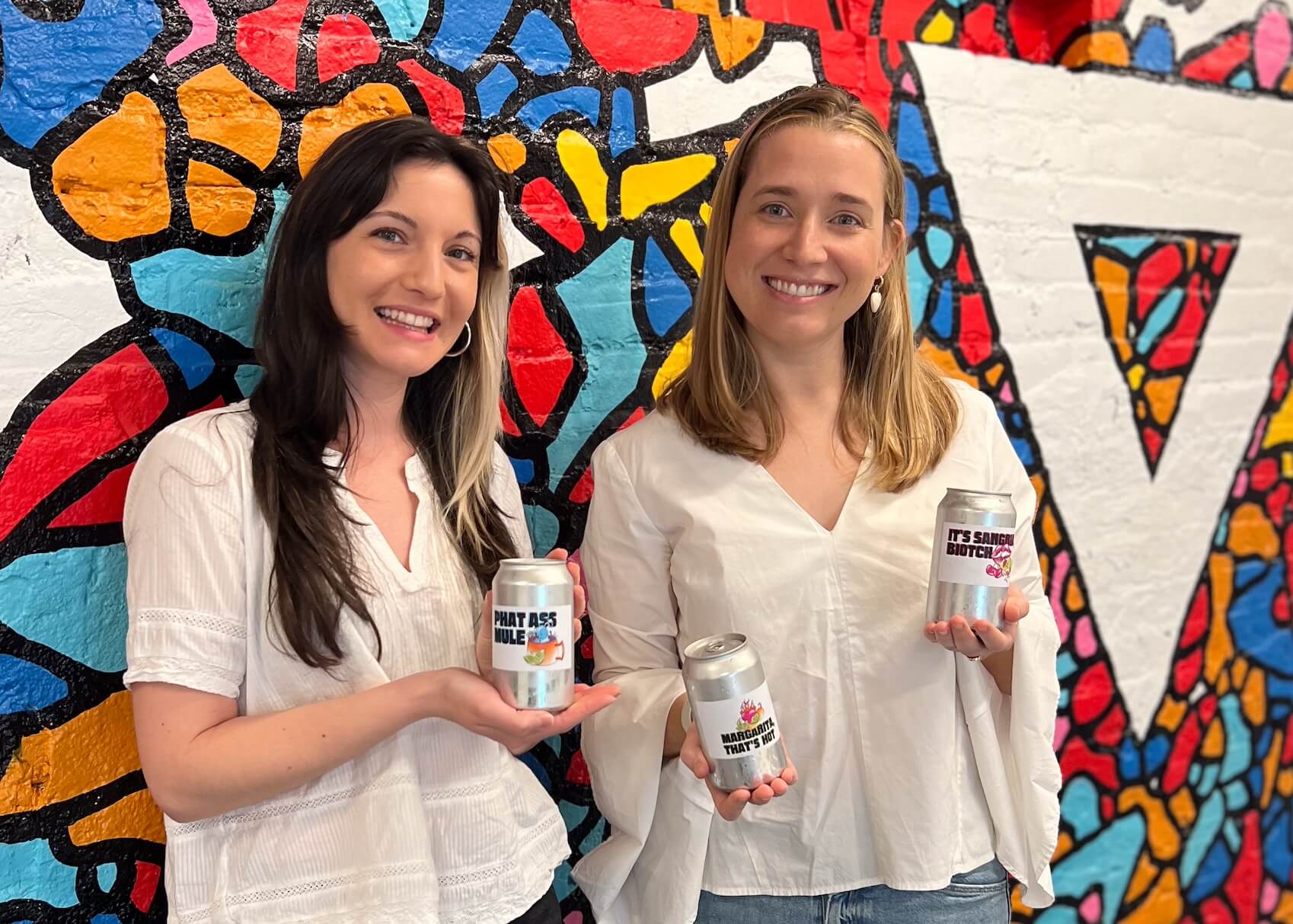 celeste and catherine holding cans of e b t b in front of the brandcenter mural in basement
